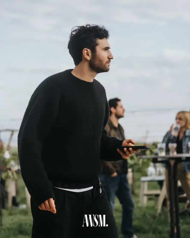 Gast im schwarzen Strickpullover mit Handy im Profil, dahinter Gäste an Stehtischen im Weinberg – Vineyard Edition, 24.05.2025
