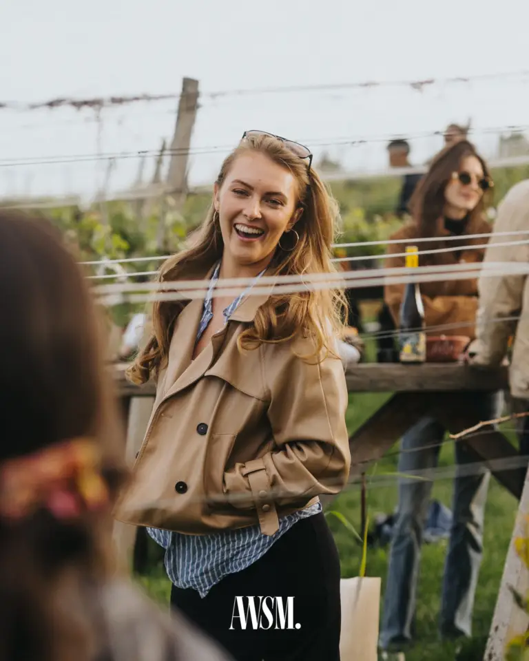 Lachender Gast in beiger Jacke an einem Tisch zwischen den Spanndrähten der Rebzeilen, dahinter Gäste – Vineyard Edition, 24.05.2025