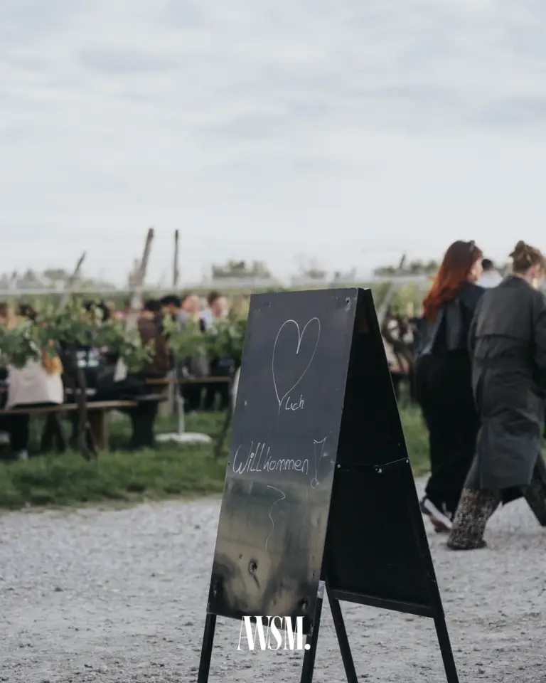 Kreidetafel mit Herz und „Willkommen“ am Kiesweg, dahinter Gäste an Tischen zwischen den Rebzeilen – Vineyard Edition, 24.05.2025