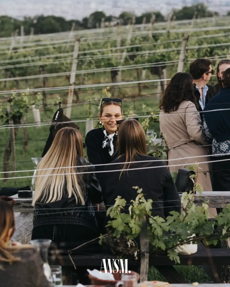 Gäste an einem Holztisch zwischen den Rebzeilen, ein Gast lächelt, dahinter der Weinberg am Nussberg – Vineyard Edition, 24.05.2025