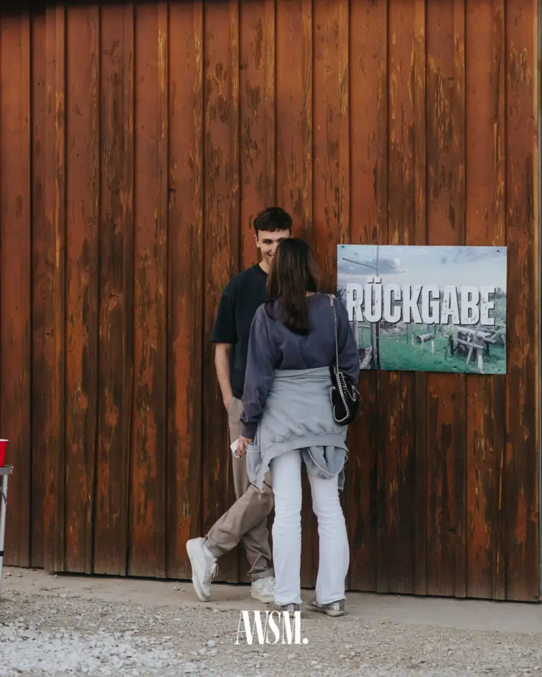Zwei Gäste stehen sich vor der Holzwand des Buschenschanks gegenüber, daneben ein Schild „Rückgabe“ – Vineyard Edition, 24.05.2025