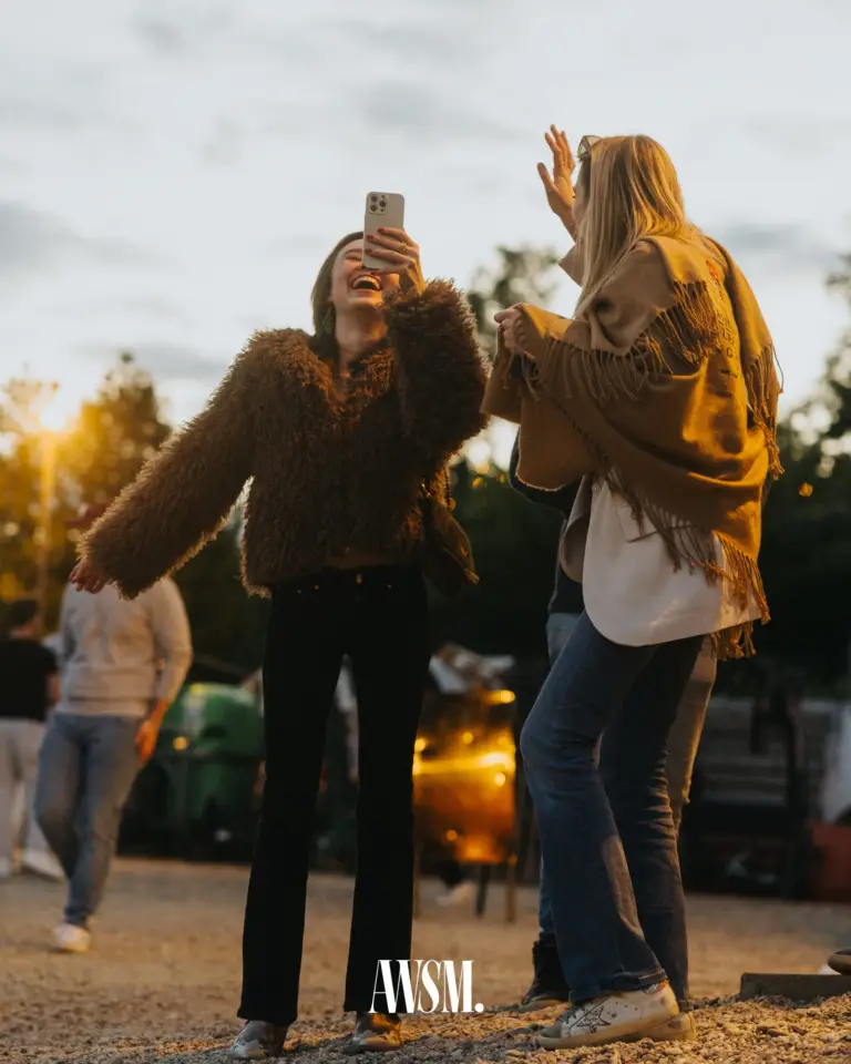 Zwei Gäste lachen und tanzen auf dem Kiesplatz, eine fotografiert mit dem Handy, dahinter die Abendsonne – Vineyard Edition, 24.05.2025