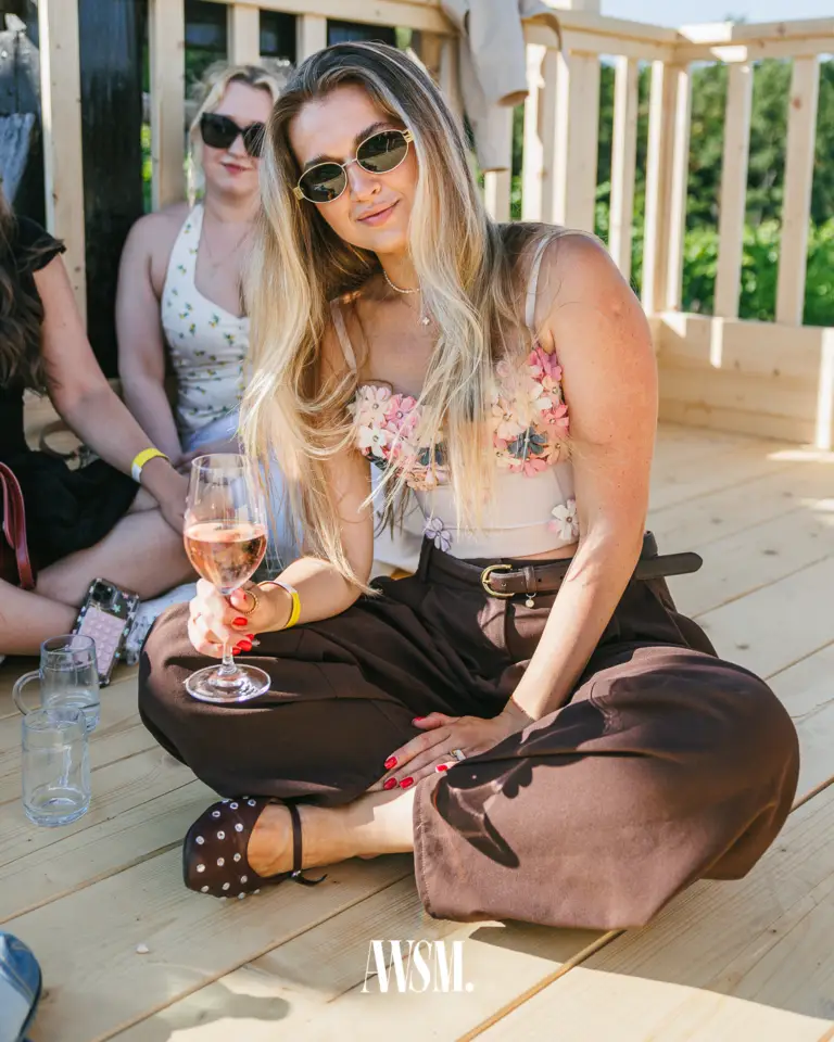 Gast mit Sonnenbrille und Glas Rosé sitzt lächelnd auf der Holzterrasse, dahinter weitere Gäste und Grün – Vineyard Edition, 19.07.2025