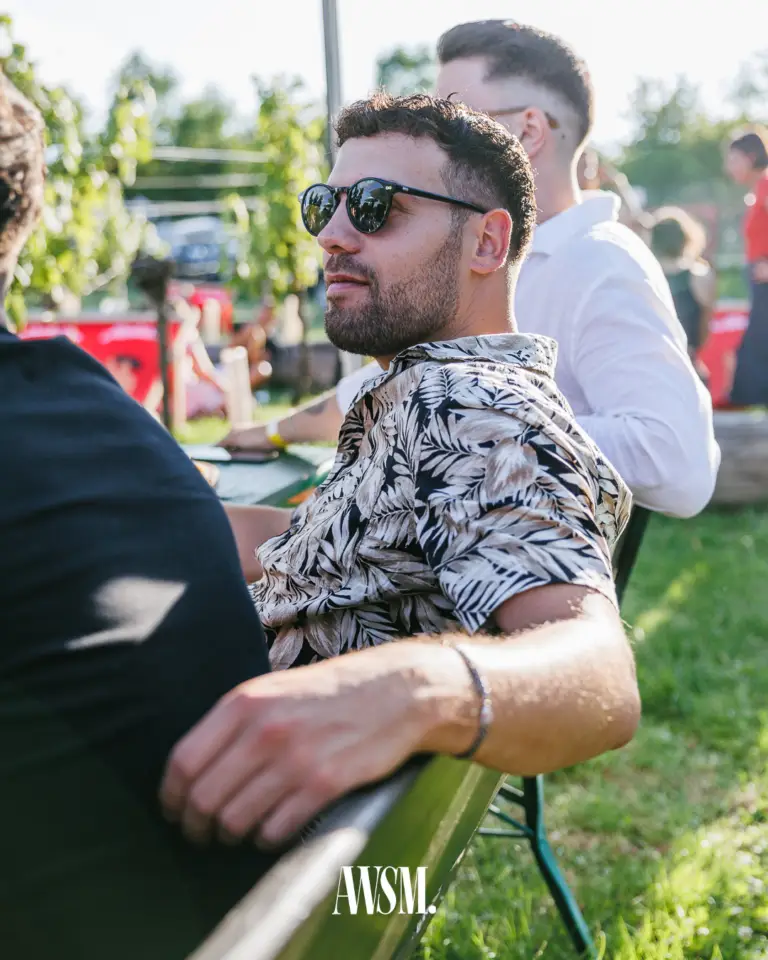 Gast mit Sonnenbrille und gemustertem Hemd sitzt auf einer Bank im Weinberg, dahinter weitere Gäste im Nachmittagslicht – Vineyard Edition, 19.07.2025