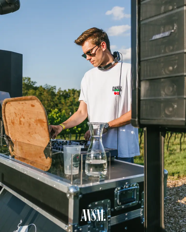 DJ mit Sonnenbrille und Kopfhörern um den Hals legt zwischen Lautsprechern auf, dahinter Weinreben und blauer Himmel – Vineyard Edition, 19.07.2025