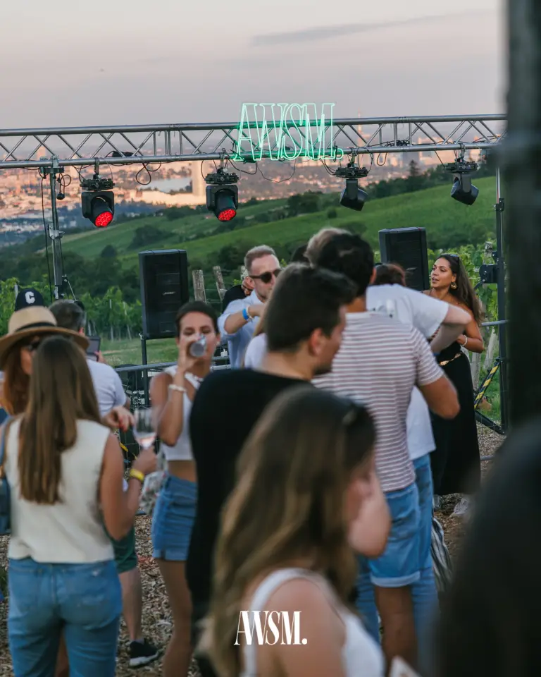 Guests celebrating in front of the lighting truss with AWSM sign and red spotlights, vineyards and city view behind at dusk – Vineyard, 19 July 2025