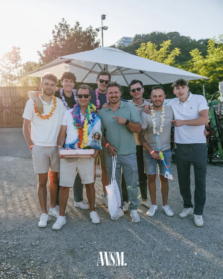 Acht Männer mit bunten Blumenketten posieren unter einem großen Sonnenschirm auf dem Kiesplatz im Gegenlicht – Vineyard Edition, 14.06.2025