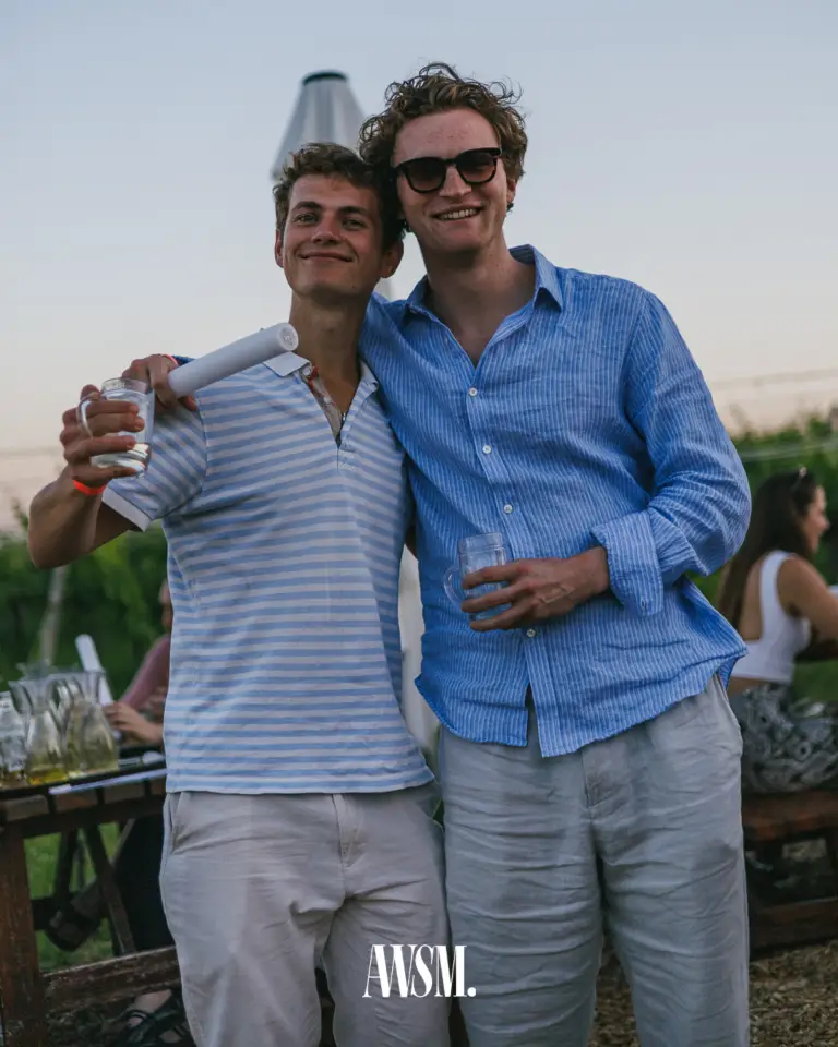 Zwei Männer in blau-weißen Hemden posieren Arm in Arm mit Gläsern und Leuchtstab vor Tischen im Weingarten – Vineyard Edition, 14.06.2025