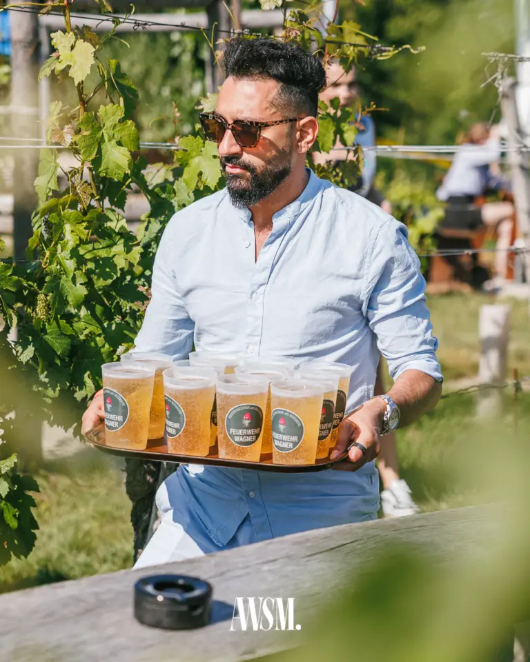 Mann mit Sonnenbrille trägt ein Tablett voller Bierbecher zwischen den Weinreben in der Sonne – Vineyard Edition, 14.06.2025