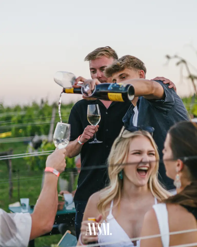Mann schenkt schwungvoll aus der Flasche in erhobene Gläser ein, davor lacht eine Frau ausgelassen – Vineyard Edition, 14.06.2025