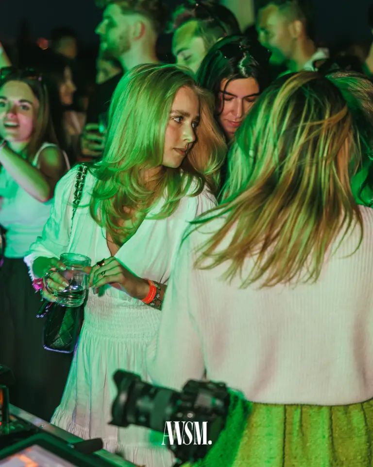 Frau im weißen Kleid mit Glas steht in grünem Licht am DJ-Pult, im Vordergrund eine Kamera, rundherum Gäste – Vineyard Edition, 14.06.2025