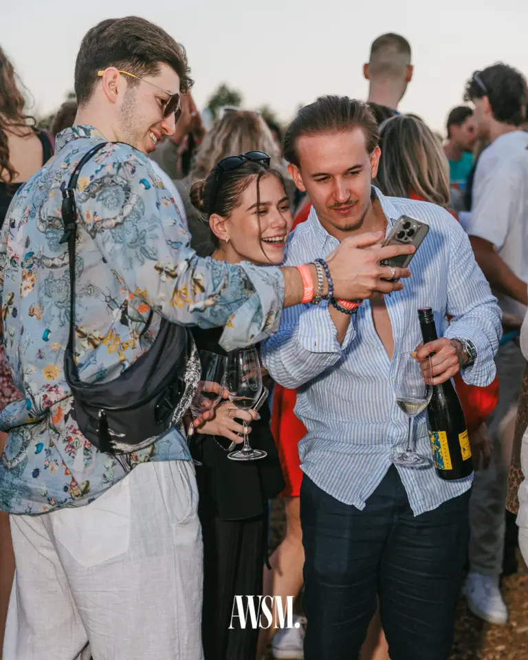 Drei Gäste schauen lachend auf ein Handy, mit Weingläsern und Weinflasche inmitten der Menge im Abendlicht – Vineyard Edition, 14.06.2025