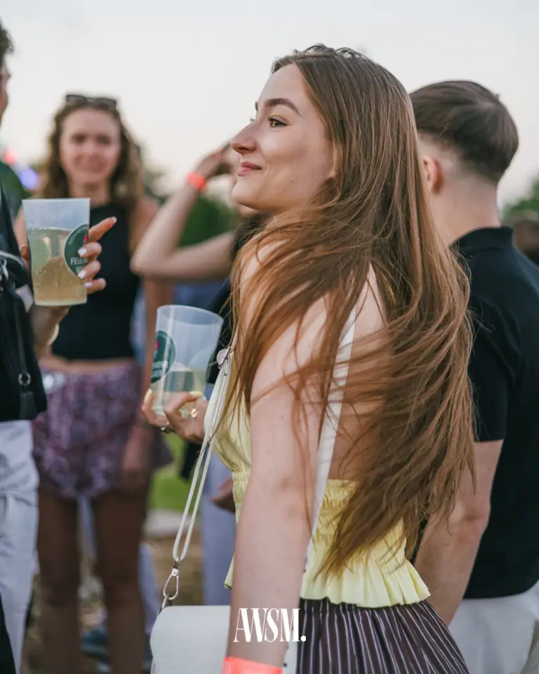Gast mit langen Haaren lächelt zur Seite, einen Becher in der Hand, weitere Gäste mit Getränken im Abendlicht – Vineyard Edition, 14.06.2025