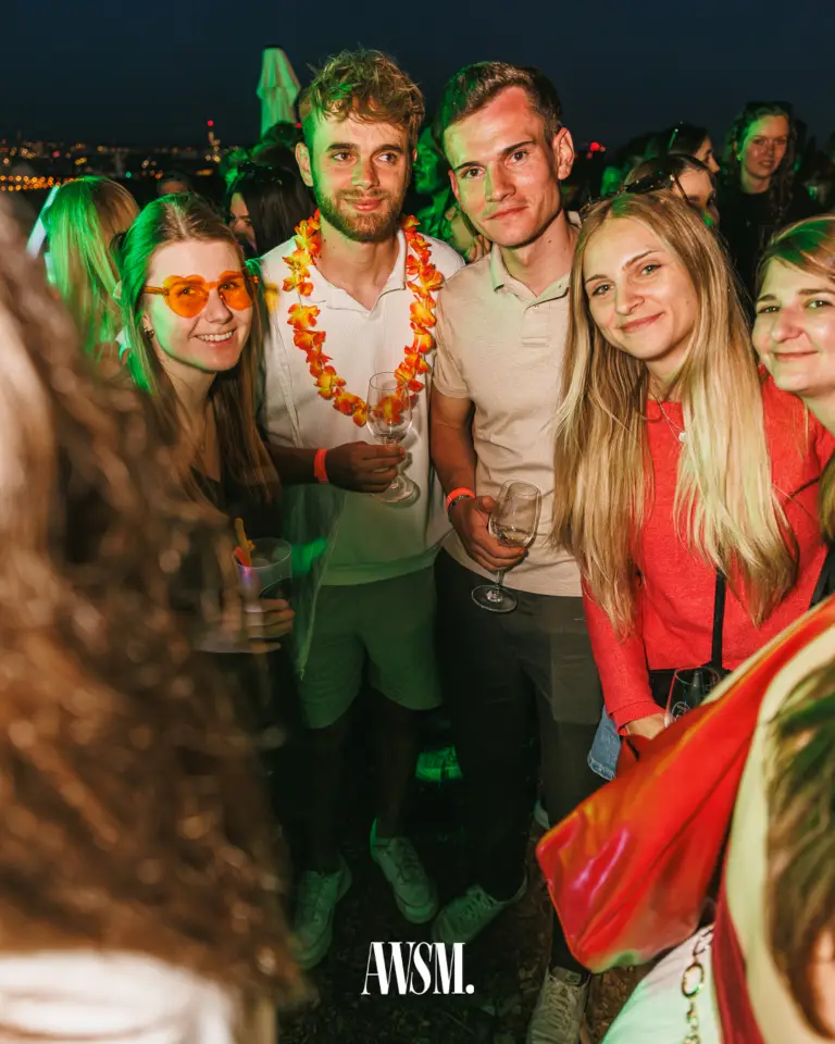Gruppe mit Weingläsern und Blumenkette posiert bei Nacht am Nussberg, im Hintergrund die Lichter der Stadt – Vineyard Edition, 14.06.2025