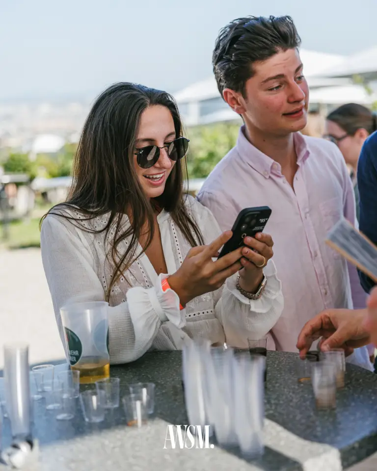 Zwei Gäste schauen am Stehtisch mit Shotgläsern und Bechern auf ein Handy, im Hintergrund der Stadtblick in der Sonne – Vineyard Edition, 14.06.2025
