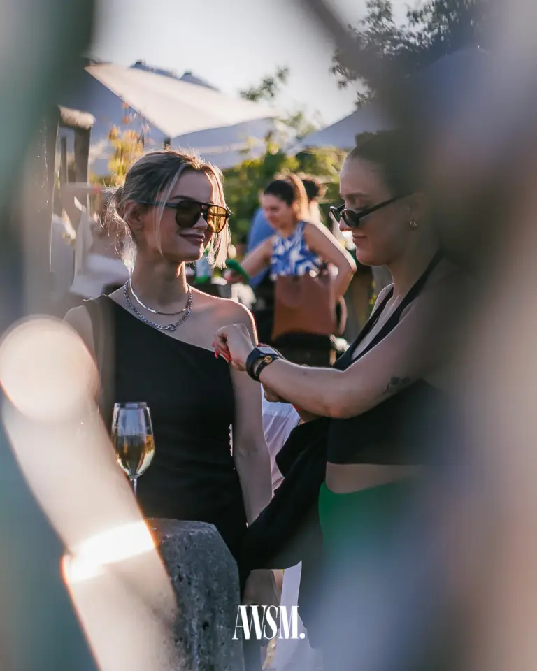 Zwei Gäste mit Sonnenbrillen stehen im Gespräch vor Sonnenschirmen; im Vordergrund ein Weinglas auf dem Tisch – Vineyard Edition, 14.06.2025