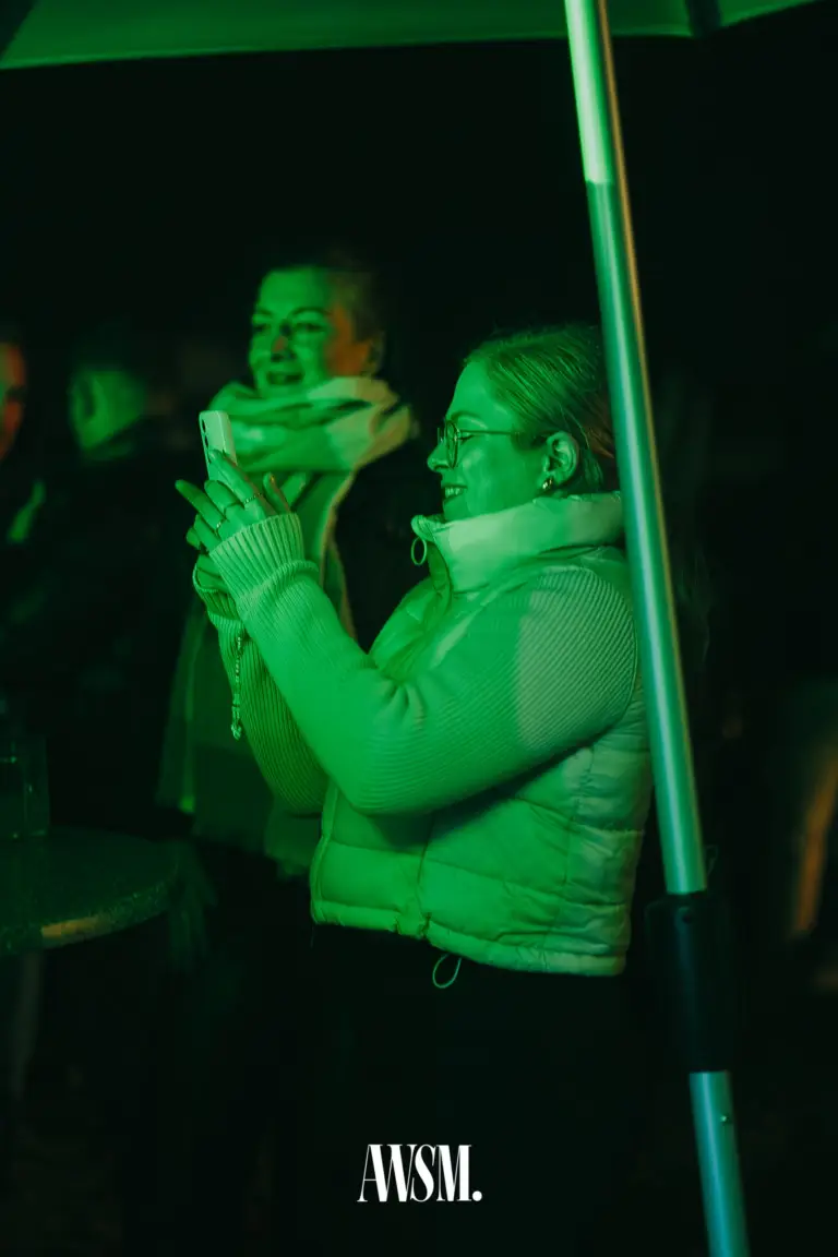Gast mit Brille und Steppweste fotografiert mit dem Handy unter grün beleuchtetem Schirm – Vineyard Edition, 04.10.2025