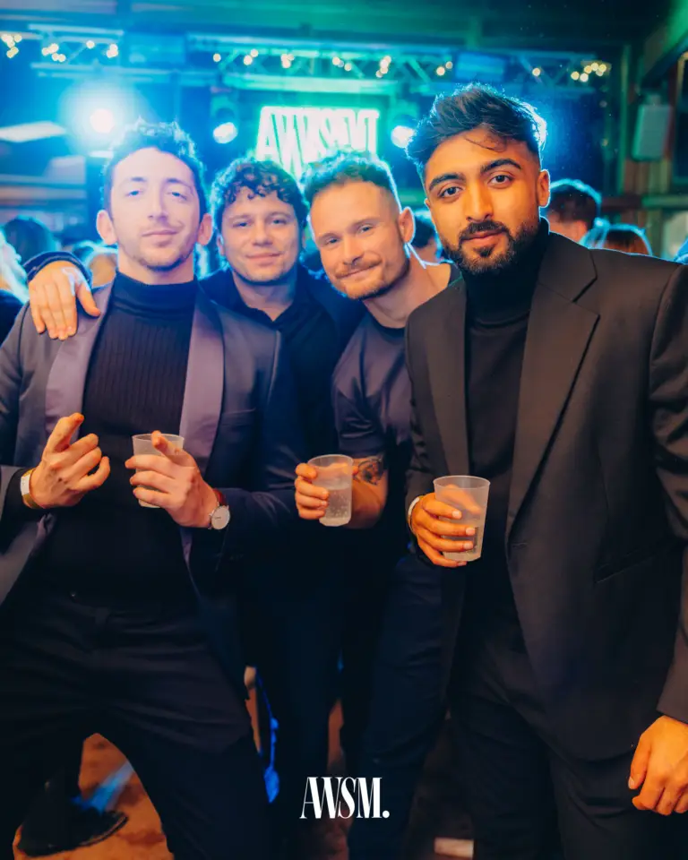 Vier Gäste in dunklen Outfits posieren mit Bechern in der Hand vor dem leuchtenden Neonlogo und Scheinwerfern – NYE Skyline Edition, 31.12.2025