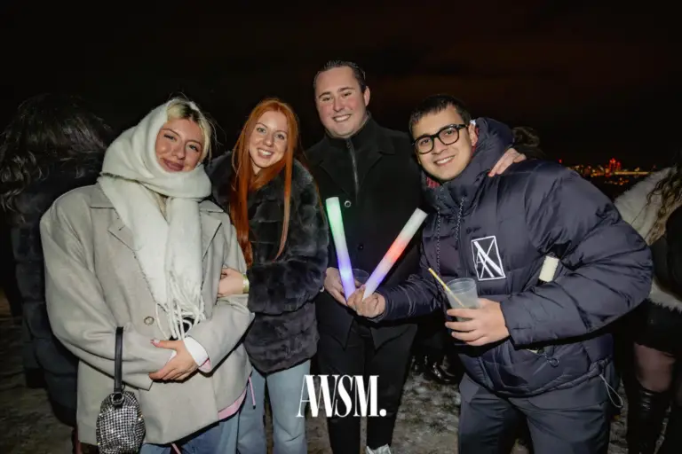 Vier Gäste in Wintermänteln posieren draußen in der Nacht mit Leuchtstäben und Bechern, hinten Stadtlichter – NYE Skyline Edition, 31.12.2025