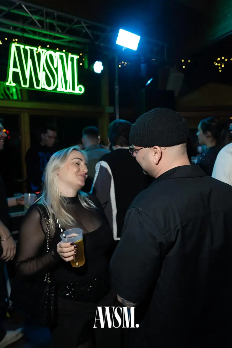 Zwei Gäste im Gespräch, einer mit Bierbecher und blonden Haaren, dahinter das grüne AWSM-Neonlogo – NYE Skyline Edition, 31.12.2025