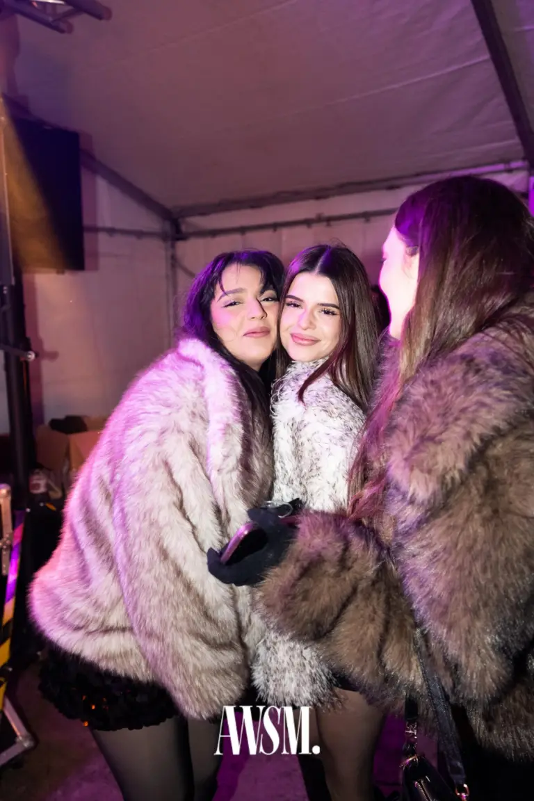 Drei Gäste in flauschigen Mänteln, zwei davon lächeln Wange an Wange im Zelt in violettem Licht – NYE Skyline Edition, 31.12.2025