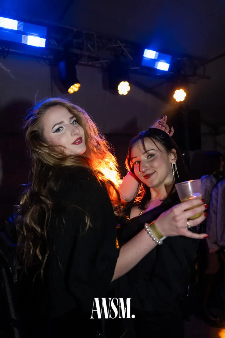 Zwei Gäste in Schwarz posieren mit einem Drink im Zelt, darüber blaue und gelbe Scheinwerfer an der Traverse – NYE Skyline Edition, 31.12.2025