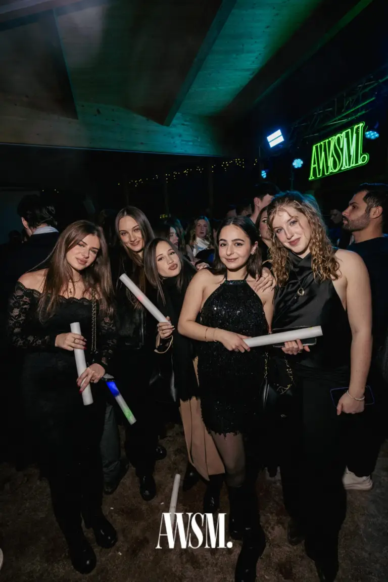 Fünf Gäste in festlichen Outfits posieren mit Leuchtstäben, dahinter grünes AWSM-Neonlogo und Holzdecke – NYE Skyline Edition, 31.12.2025
