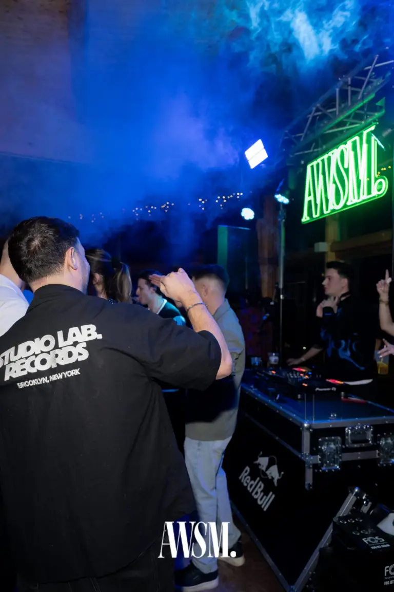 Gäste von hinten im blauen Nebel vor dem DJ-Pult, darüber das grün leuchtende AWSM-Neonlogo an der Traverse – NYE Skyline Edition, 31.12.2025