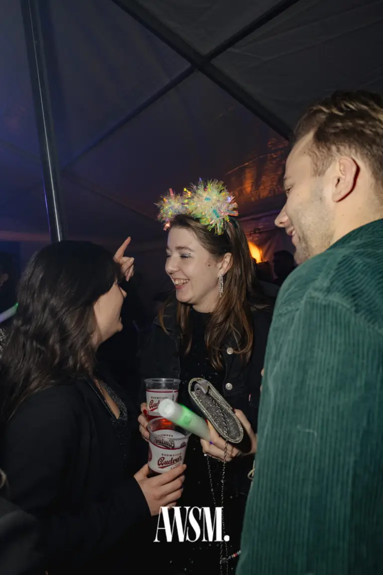 Gast mit glitzerndem Lametta-Haarschmuck lacht mit Bechern und Leuchtstab in der Hand, im Zelt mit Gästen – NYE Skyline Edition, 31.12.2025