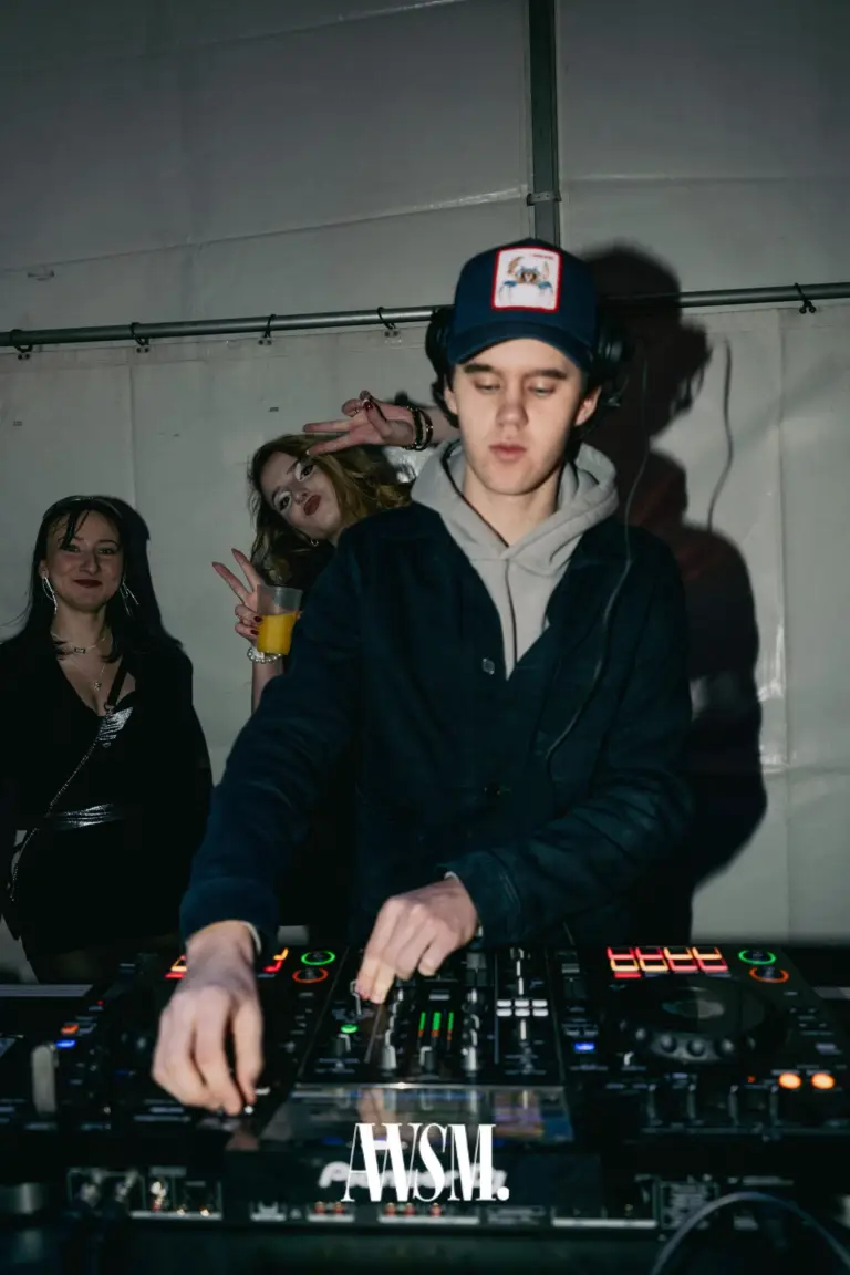 DJ mit Kappe und Kopfhörern mischt im Zelt, hinter ihm posieren Gäste mit Drink und Peace-Zeichen – NYE Skyline Edition, 31.12.2025