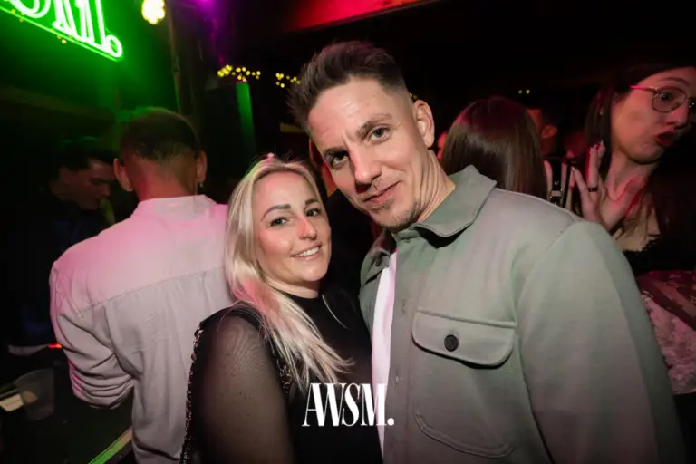 Zwei Gäste posieren eng beisammen und lächeln in die Kamera, dahinter Gäste in pinkem Licht und Neonschrift – NYE Skyline Edition, 31.12.2025