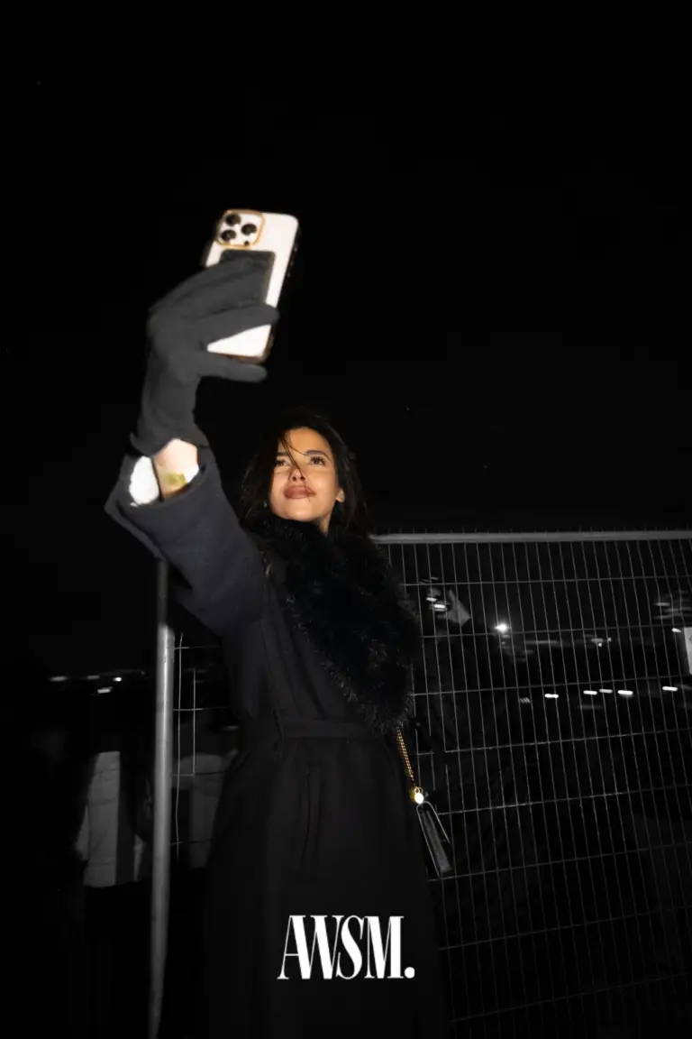 Gast in schwarzem Mantel mit Pelzkragen und Handschuhen macht nachts draußen ein Selfie vor einem Gitterzaun – NYE Skyline Edition, 31.12.2025