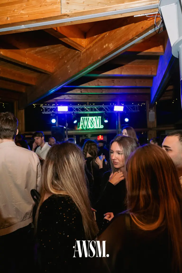 Gäste im Gespräch unter der hölzernen Dachkonstruktion, im Hintergrund die Bühne mit grünem AWSM-Neonlogo – NYE Skyline Edition, 31.12.2025