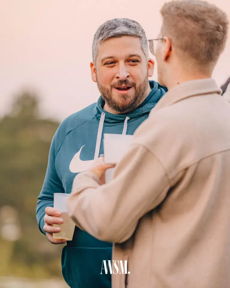 Mann mit grauem Haar und Bart im Kapuzenpulli im Gespräch mit einem Gast, beide mit Bechern, dahinter Abendhimmel – Grand Vineyard Opening, 11.04.2026