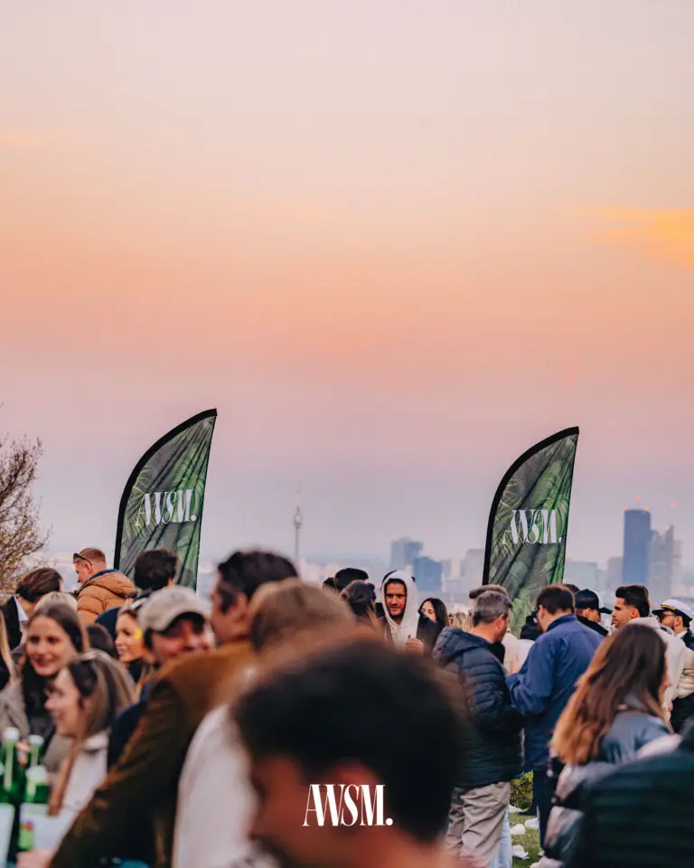 Gäste feiern im Abendrot am Reisenberg zwischen AWSM-Fahnen, dahinter die Skyline von Wien – Grand Vineyard Opening, 11.04.2026