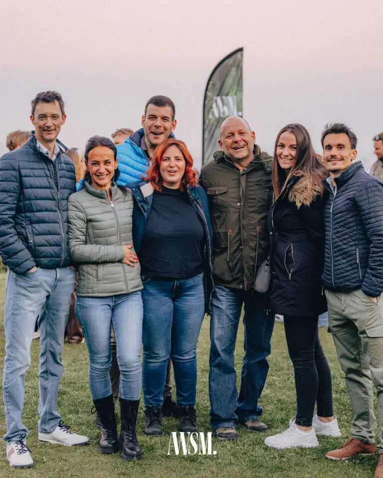 Gruppe lachender Gäste in Jacken posiert auf der Wiese, dahinter eine Beachflag und blasser Abendhimmel – Grand Vineyard Opening, 11.04.2026