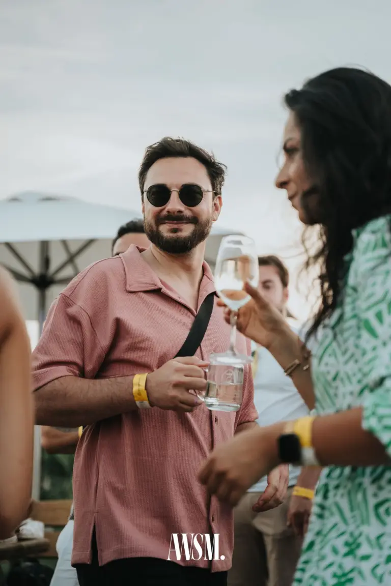 Gast mit runder Sonnenbrille und rosa Hemd hält ein Glas, daneben Frau mit Weinglas unter Schirmen am Nussberg – Vineyard Summer Opening, 20.06.2026