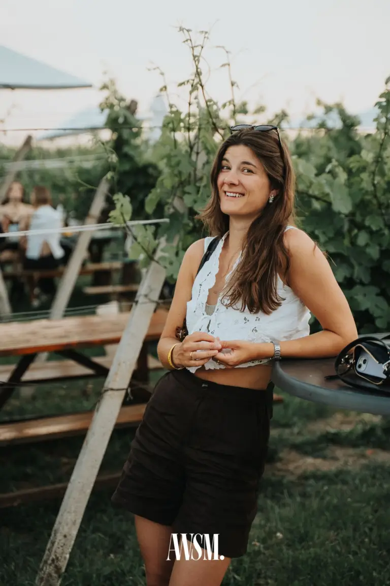Lächelnde Frau in weißer Spitzenbluse lehnt an einem Stehtisch zwischen Weinreben und Holzbänken am Nussberg – Vineyard Summer Opening, 20.06.2026