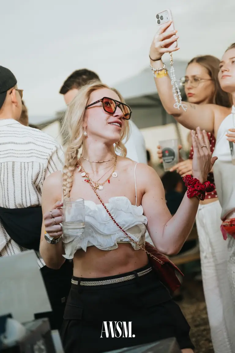 Blonde Frau mit Zopf und getönter Sonnenbrille hält ein Glas, hinter ihr filmt ein Gast mit dem Handy am Nussberg – 20.06.2026