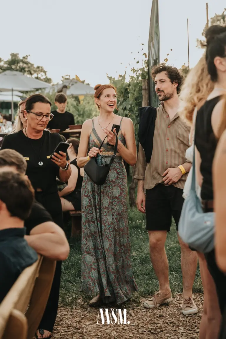 Lachende Frau im gemusterten Maxikleid mit Handtasche neben Gast im Streifenhemd zwischen Weinreben am Nussberg – Vineyard Summer Opening, 20.06.2026