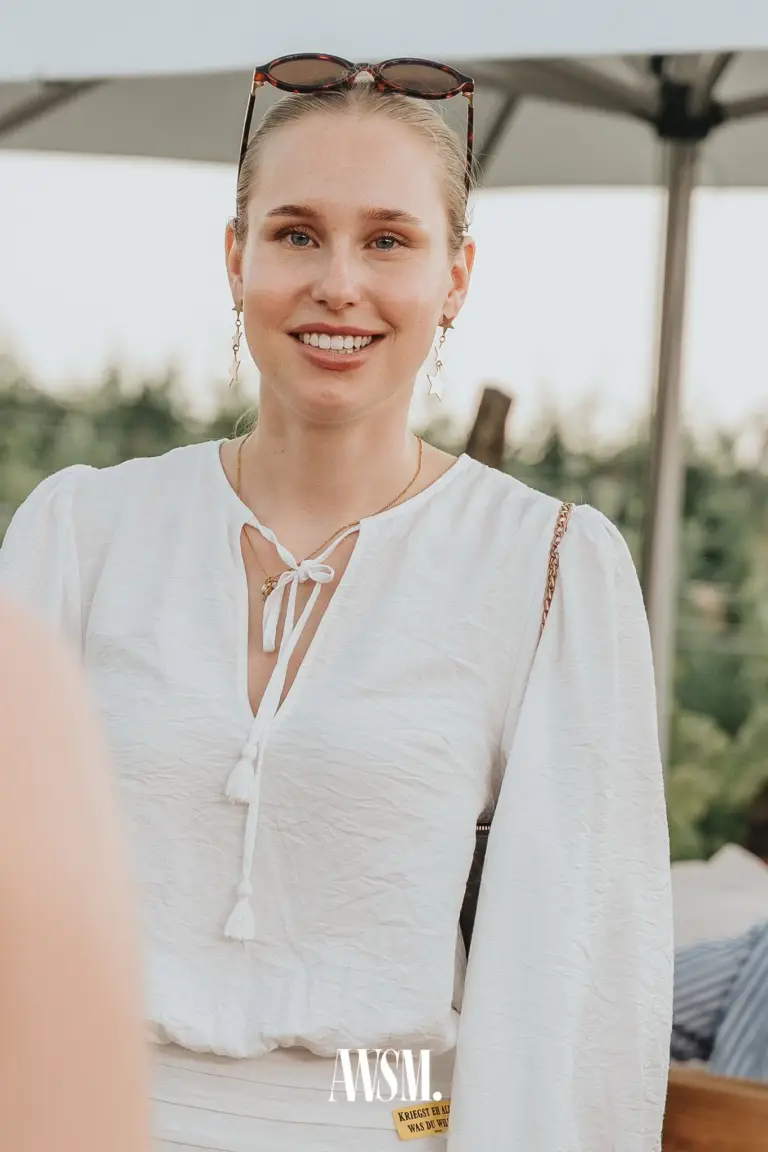 Lächelnde Frau in weißer Bluse mit Sonnenbrille im Haar unter einem Schirm vor dem Weinberg am Nussberg – Vineyard Summer Opening, 20.06.2026