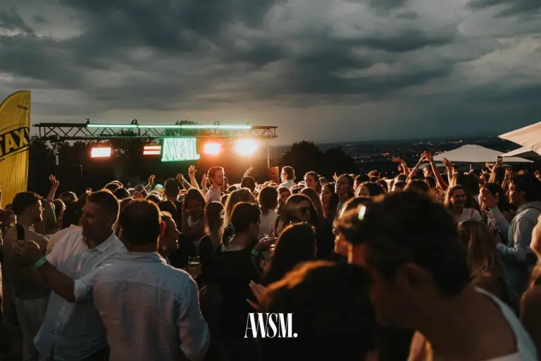 The crowd parties in front of the stage with the AWSM neon sign and spotlights, dark clouds above, a city view on the right – 18 July 2026