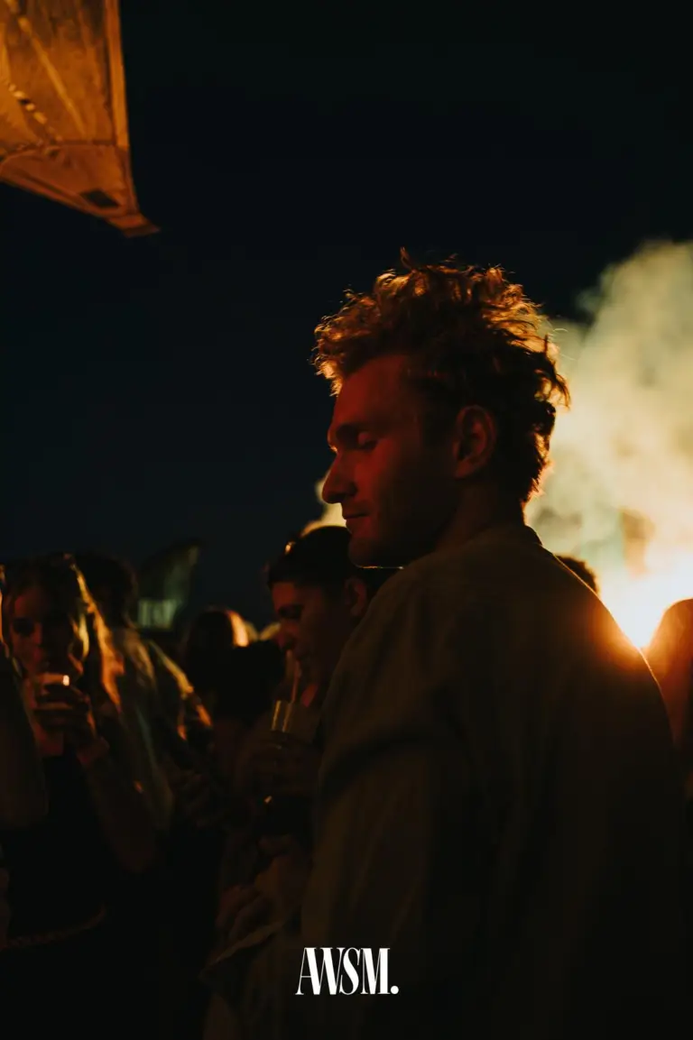 Mann mit lockigem Haar im Profil vor leuchtendem Nebel bei Nacht, daneben Gäste mit Getränken – Skyline Edition, 08.08.2026
