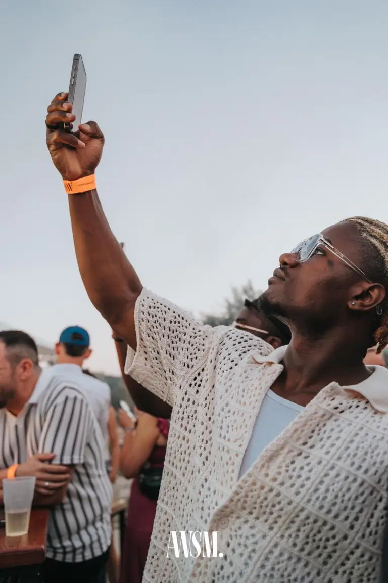 Mann im Häkelhemd mit heller Sonnenbrille hält sein Handy in den Himmel, dahinter Gäste an einem Tisch – Skyline Edition, 08.08.2026