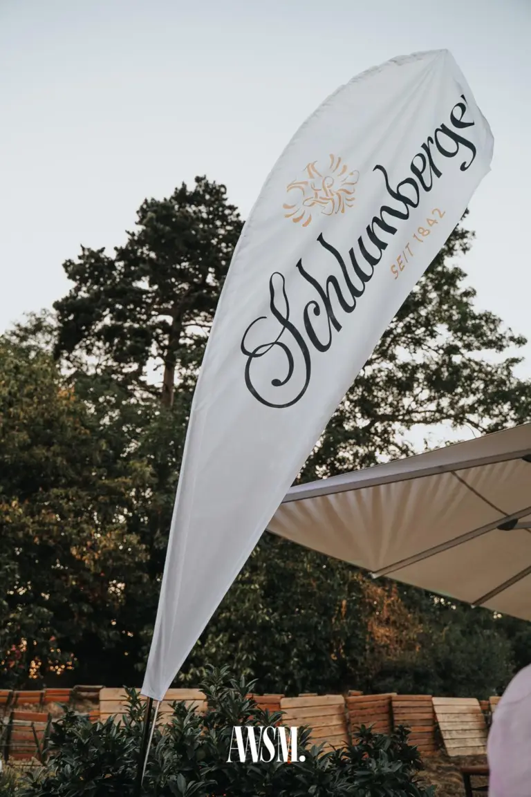 Weiße Beachflag mit Schriftzug vor hohen Bäumen, daneben ein Sonnenschirm und gestapelte Holzliegen – Skyline Edition, 08.08.2026