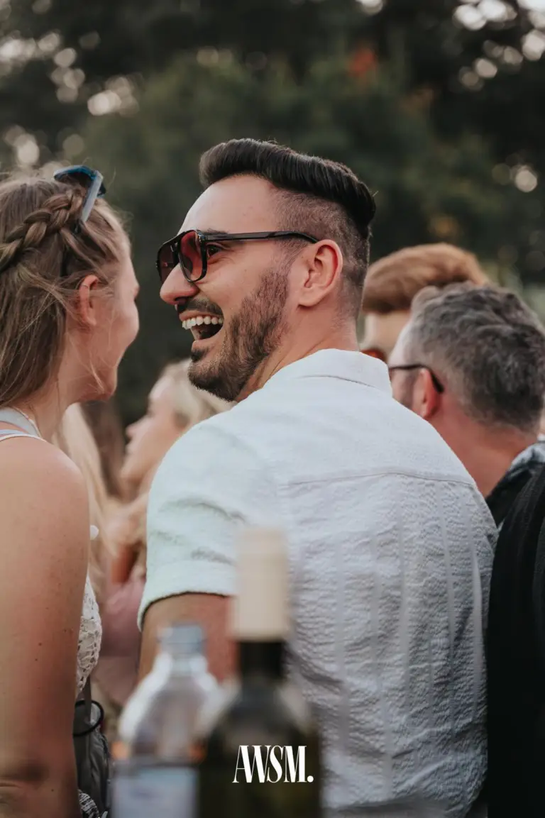 Bärtiger Mann mit Sonnenbrille lacht einer Frau mit Flechtfrisur zu, vorne unscharf Flaschen, dahinter Bäume – Skyline Edition, 08.08.2026