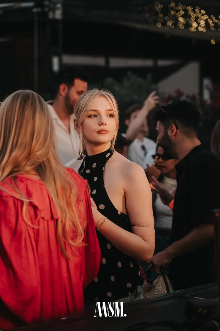 Blonde Frau im gepunkteten Neckholder-Top blickt zur Seite, vorne eine Frau in roter Bluse, oben Lichterketten – Skyline Edition, 08.08.2026