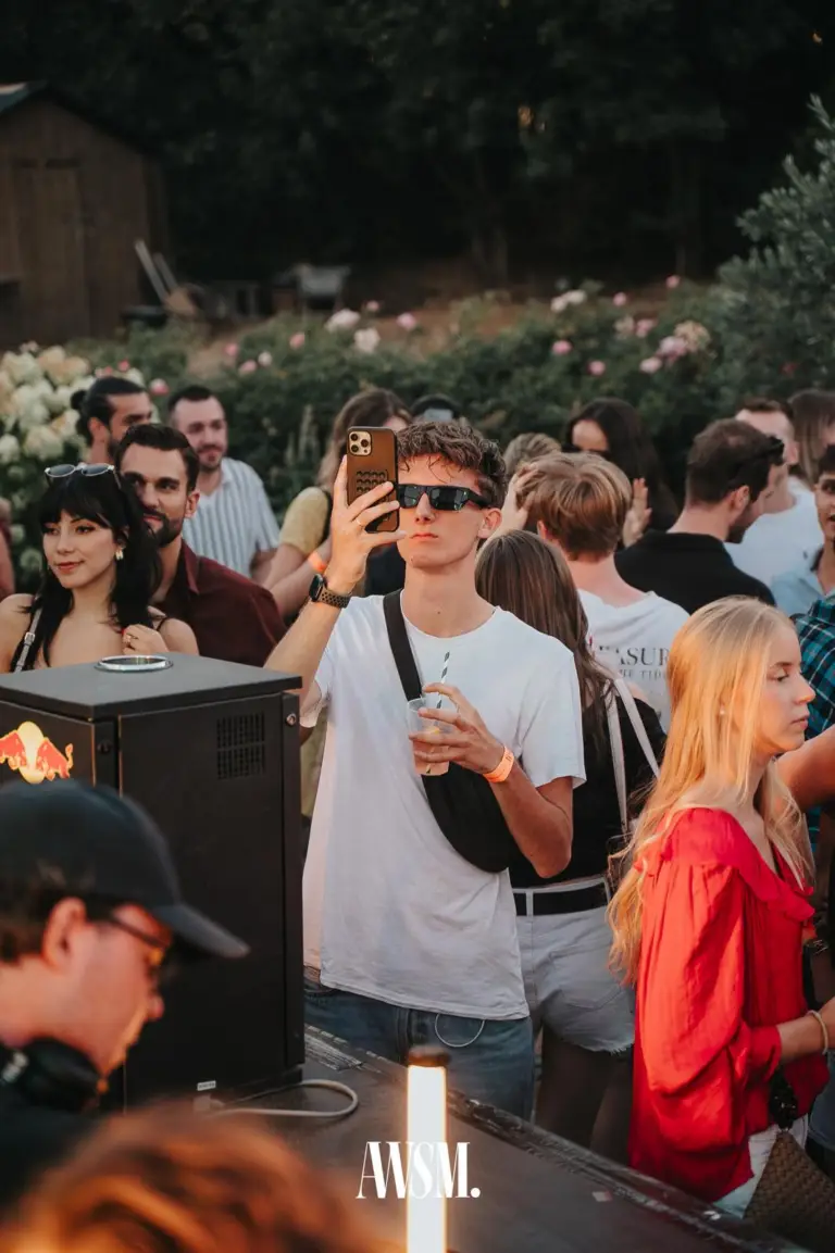 Mann mit Sonnenbrille filmt am DJ-Pult mit dem Handy, dahinter Gäste, blühende Sträucher und eine Holzhütte – Skyline Edition, 08.08.2026