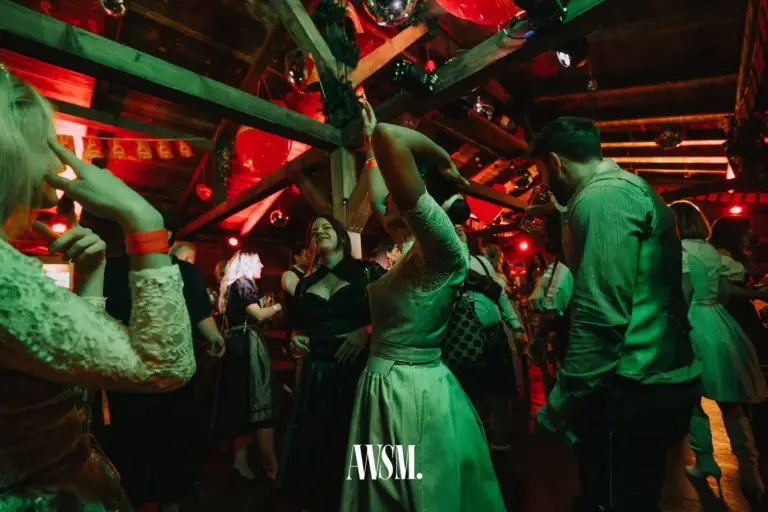 Gäste im Dirndl tanzen in grünem und rotem Licht unter Holzbalken mit Discokugeln, eine Frau hebt den Arm – AWSM. GOES KAISER WIESN, 27.09.2026