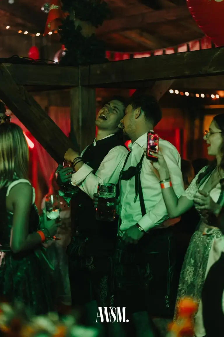 Zwei Männer in Lederhose mit Hosenträgern lachen mit Bierkrug, daneben filmt eine Frau im Dirndl mit dem Handy – AWSM. GOES KAISER WIESN, 27.09.2026